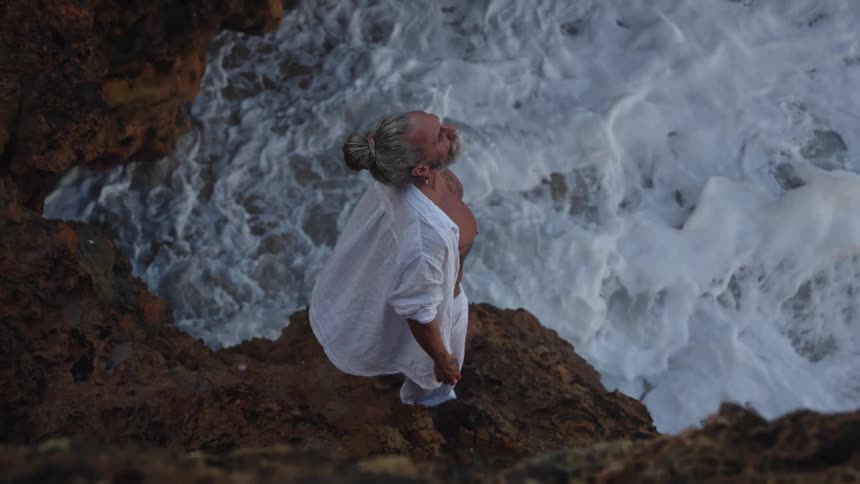 A man with a gray beard and his hair tied back stands on dark rocks at a cliffs edge looking up with his torso bare and a white shirt open while white ocean waves crash immediately below him