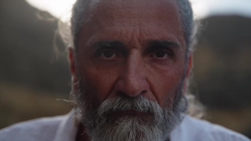 A close view shows an older man with a gray beard and gray hair tied back wearing a white shirt with his eyes opening to look directly at the camera in soft light
