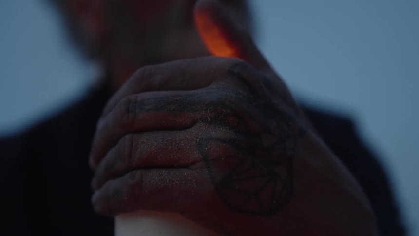 A close view shows a mans sandy tattooed hand cupping a lit white candle before the flame is blown out against a dark blue background