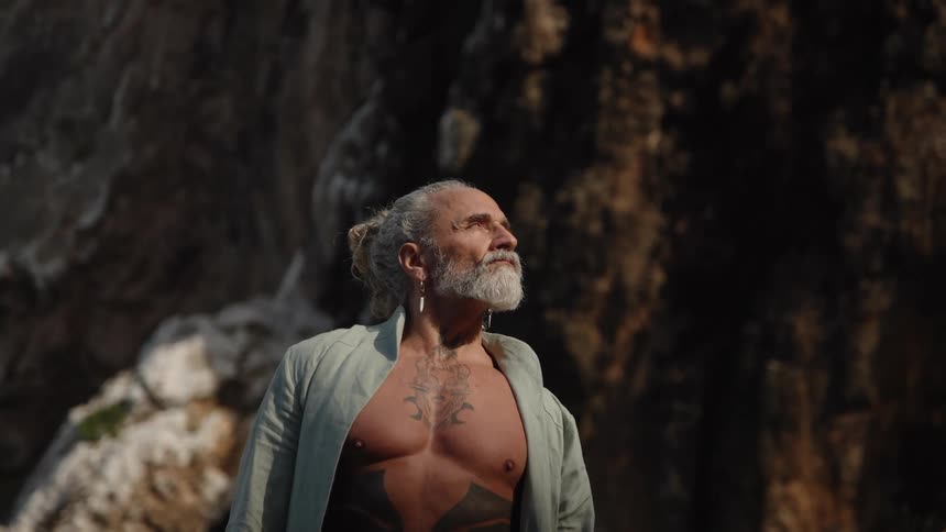 A medium view shows a shirtless muscular older man with a gray beard and hair tied back wearing an open gray shirt looking up toward the light in front of a dark textured rock wall