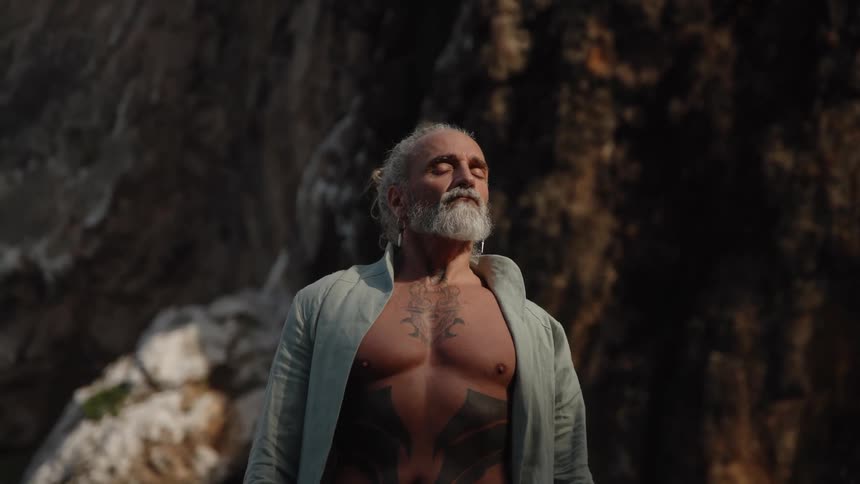 A muscular man with a gray beard and tattooed torso stands with an open jacket before a dark rock wall looking upward with a closedeye expression before lowering his gaze to look forward