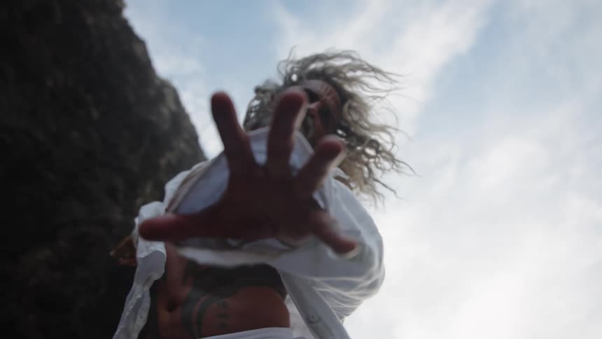 A muscular man with long windblown hair and tattoos on his chest reaches a hand out toward the camera from a low angle throwing his head back as he stands near a dark cliff