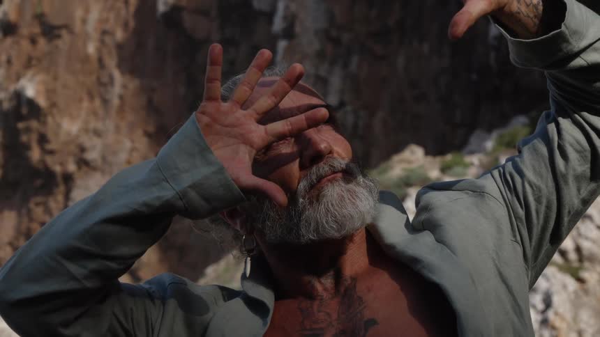 An older man with a gray beard and open jacket stands before a rock wall looking up toward the light while raising his hands to shield his eyes