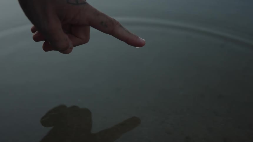 A close view shows a tattooed hand with an index finger dipping into the calm dark water creating ripples and drops of water