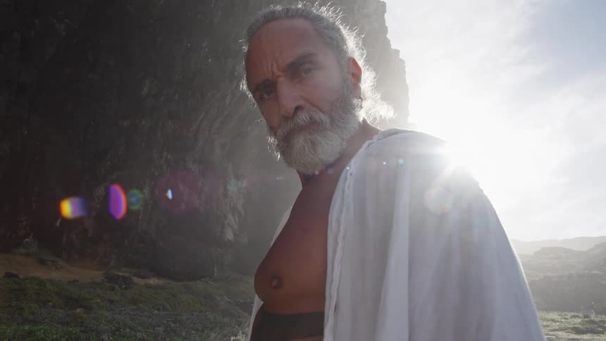 A person with facial hair and long hair stands in an outdoor area wears a white cloth over one shoulder with a large rock structure looms behind him and sunlight creates a strong outline around the head and body