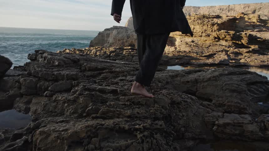 Person in dark clothing stands barefoot on rocks facing the sea with gentle waves and sunlit cliff in the background