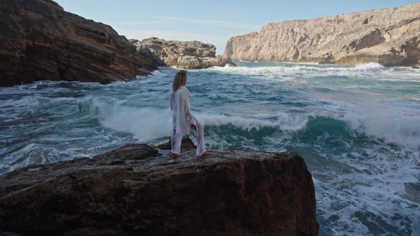 A man wearing light clothing stands on a dark rock outcrop at the edge of the sea facing the large blue waves that crash violently between steep coastal cliffs before raising his arms