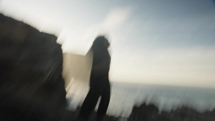 An extreme out of focus shows a man in a long black coat standing on a rocky cliff gesturing with his arms reaching up against the bright hazy sky and the ocean below