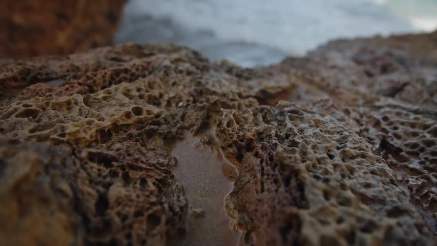 A low angle close view shows the rough porous texture of brown volcanic rock with a small pool of water in a crevice and blurry ocean waves in the background