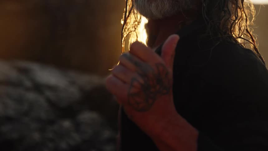 A close low angle view shows a shirtless tattooed older man with wet hair and a gray beard holding his chest with a tattooed hand as he is brightly silhouetted by the intense setting sun
