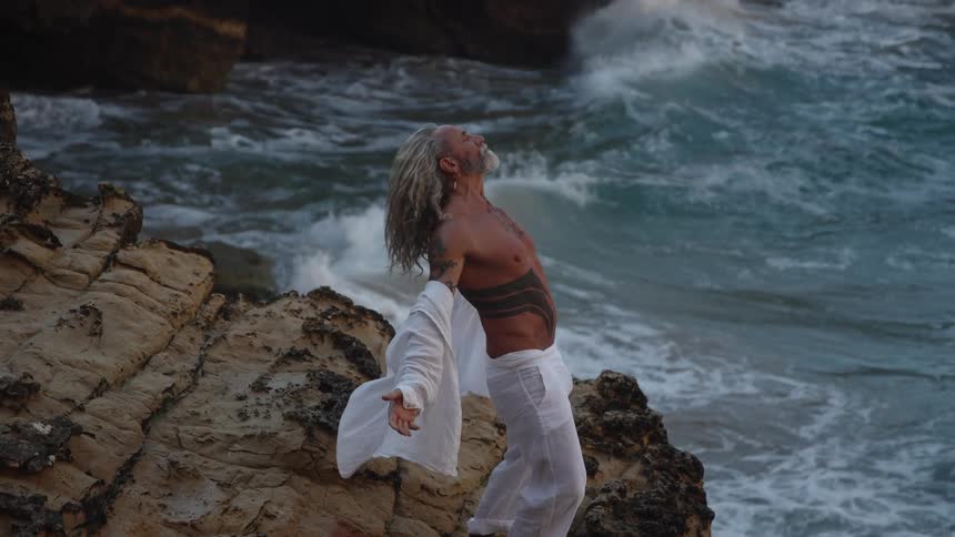 Grayhaired man with chest tattoo stands with arms outstretched on rocks with waves in the background