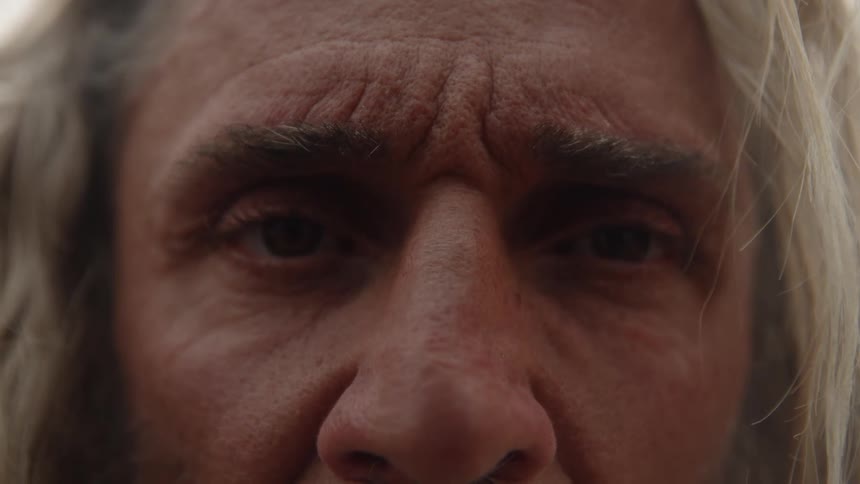 A closeup shot focuses on the eyes and forehead of a person with long gray hair who stares intently toward the viewer with a neutral expression