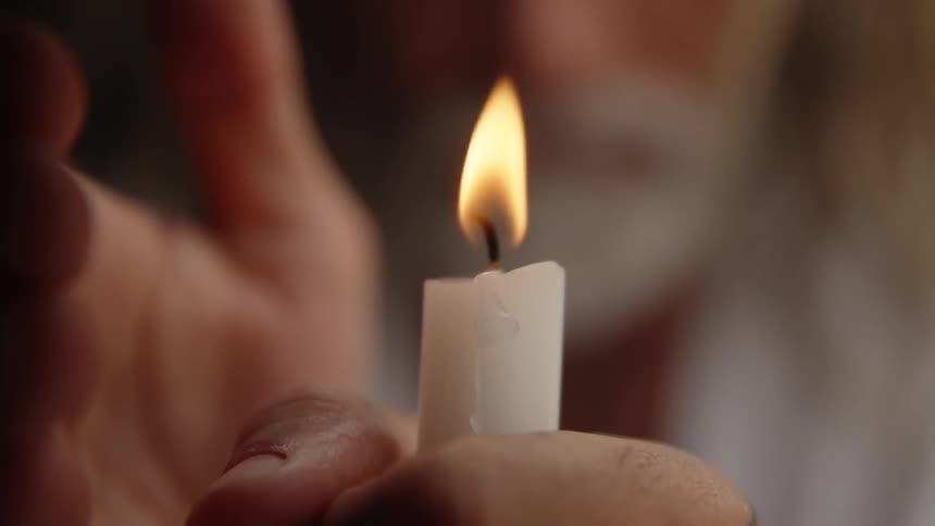 Hand holds lit white candle with steady flame in front of blurred figure in background