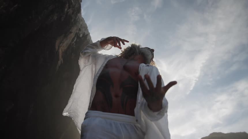 The video shows an intense lowangle closeup of a longhaired man in a white open shirt reaching his arm toward the camera while standing near a rocky cliff face outdoors the sunlight is bright behind him creating a dramatic ethereal feel as he r