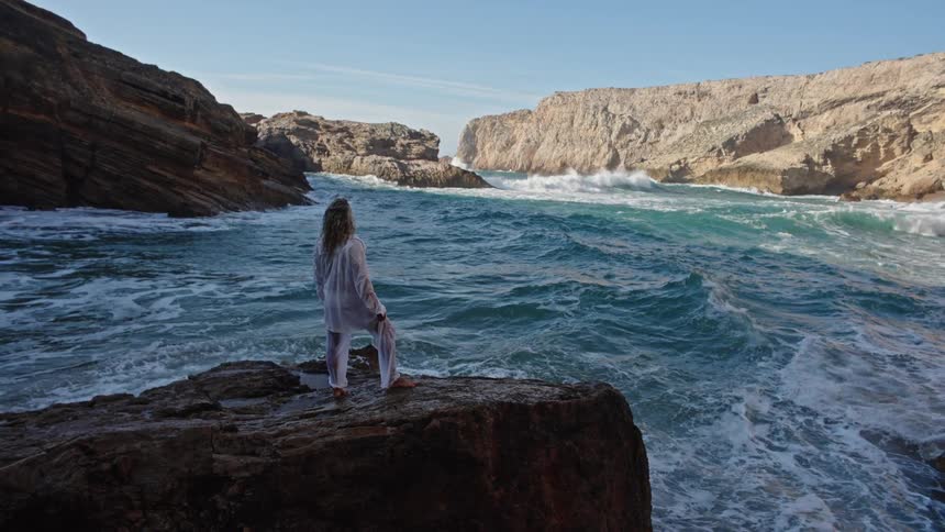 The video shows a man standing on a rugged dark cliff edge overlooking a churning deep blue sea he is facing the ocean and is wearing a flowing white sheer garment as large waves crash against the high cliffs in the distance