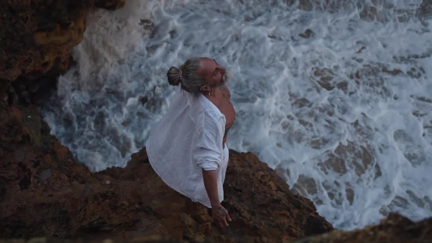 A high angle medium view shows an older man with a gray beard and hair tied in a bun wearing a white shirt and pants standing on dark rocks above the crashing ocean and lifting his arms to the sky