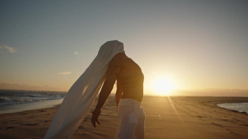 Man in motion on sand with flowing fabric at sunset by the sea