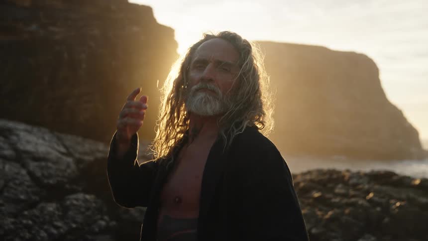 A long haired man with a gray beard and an open dark shirt stands on a rocky beach running his hand through his hair while bright sunlight flares behind him and illuminates the cliffs