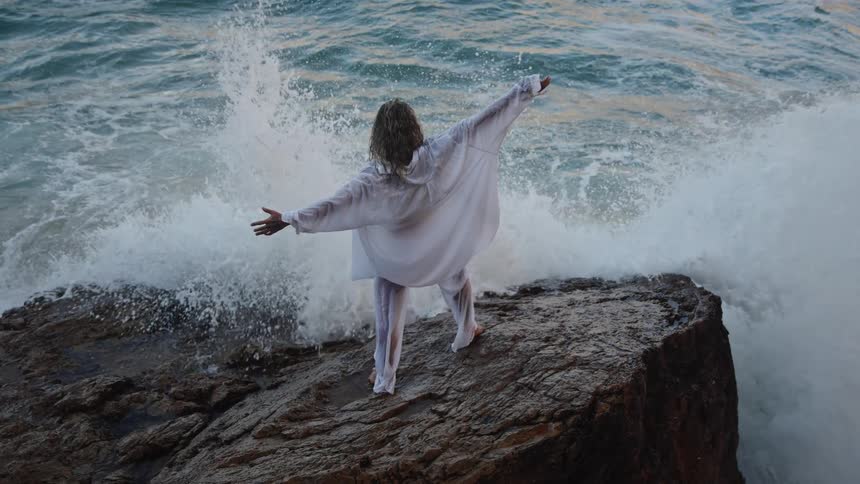 Man with arms outstretched on rocks facing the rough sea