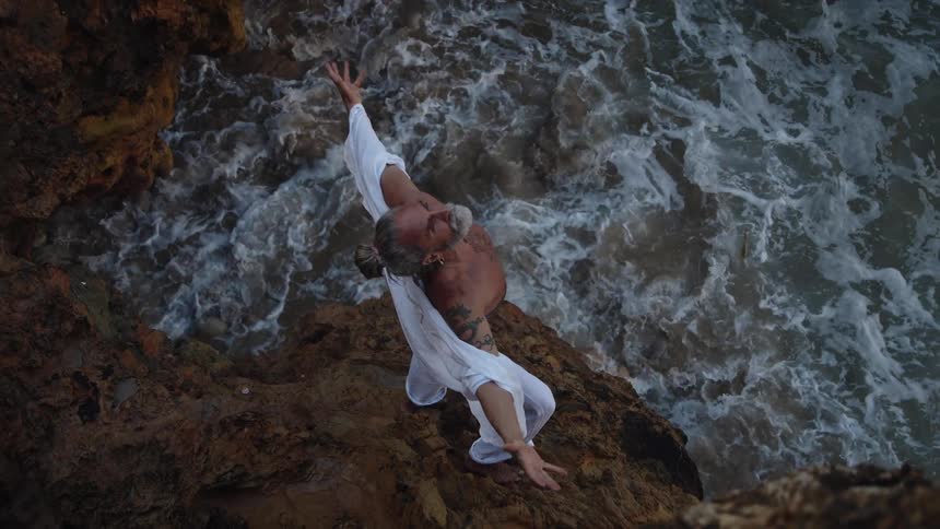Person standing with arms outstretched on rocks as waves crash below