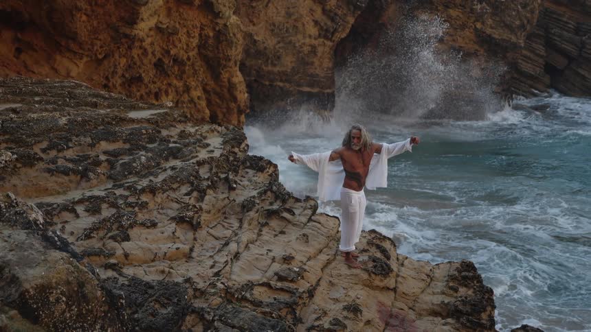 A person with long white hair and beard stands on a rugged rocky cliff overlooking the ocean wearing a white flowing open shirt and white pants large waves crash dramatically behind them spraying white foam high into the air