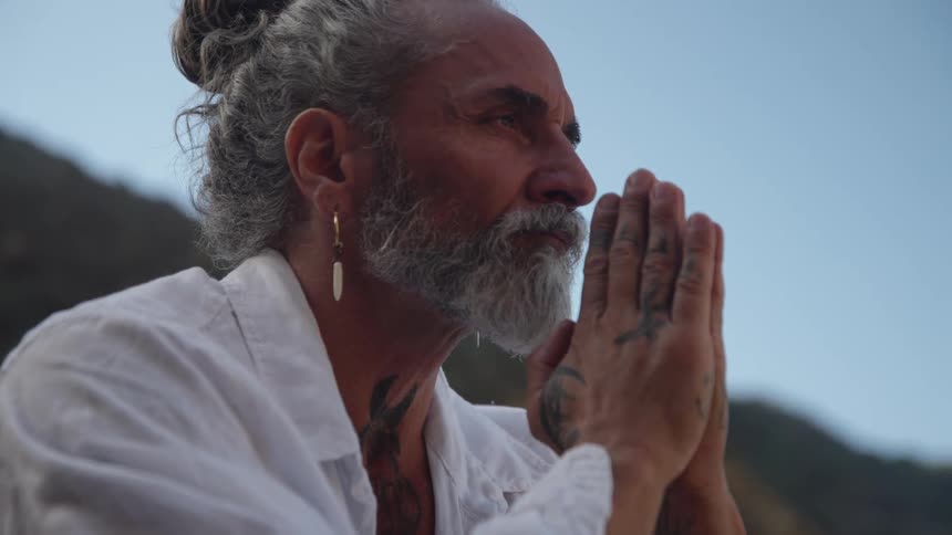 A middleaged man with gray hair and a beard wearing a white shirt sits outdoors at midday with his hands in prayer tattoos visible on his body