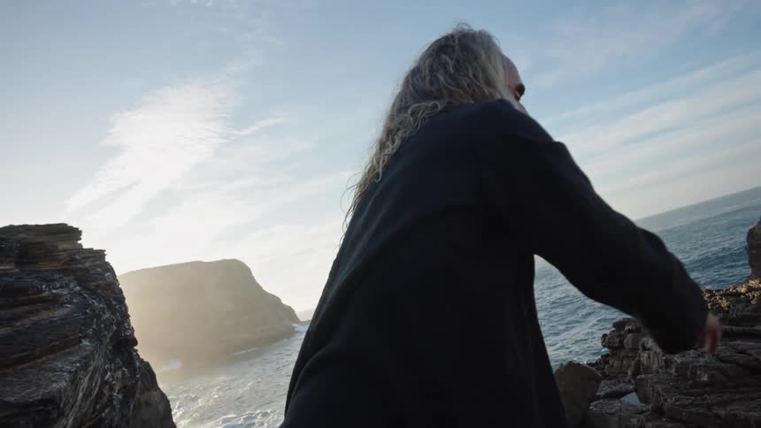 A middleaged man with a gray beard and long blond hair dances on a cliff by the shoreline at midday
