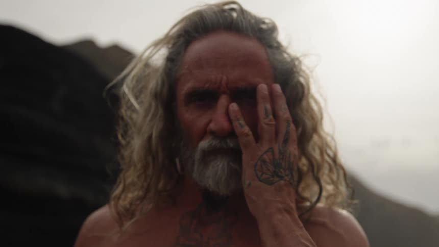 Longhaired bearded man covering one eye with his tattooed hand under soft light