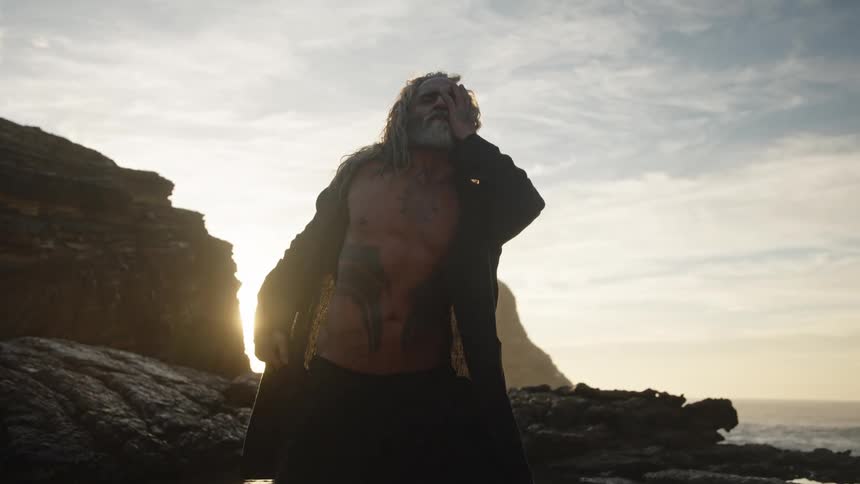 Shirtless man posing confidently on a rocky beach at sunrise