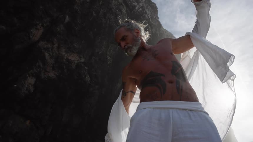 An older man with a muscular tattooed torso and gray beard moves dynamically at the base of a dark cliff spreading a white garment and reaching a hand toward the camera against a bright sun that causes a lens flare
