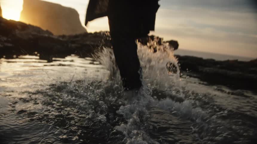 A low angle close view of a person wearing dark pants and a jacket as they wade barefoot through shallow water on a rocky beach with the setting sun creating a bright silhouette