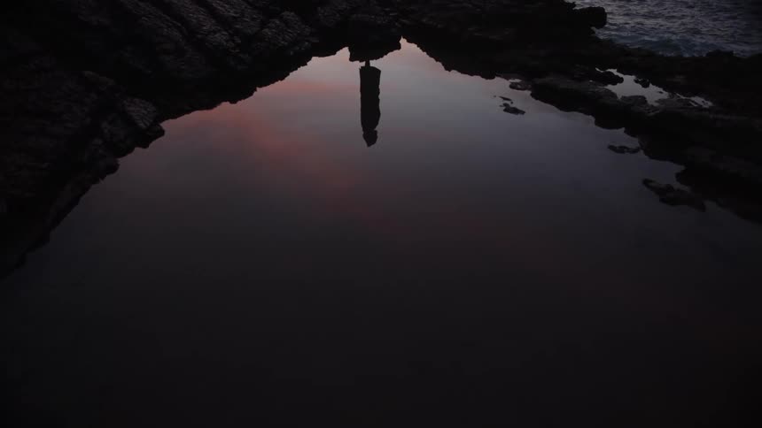 A medium view of a dark tide pool surrounded by dark rocks shows the reflection of a person standing on the edge against the subtle colors of a twilight sky