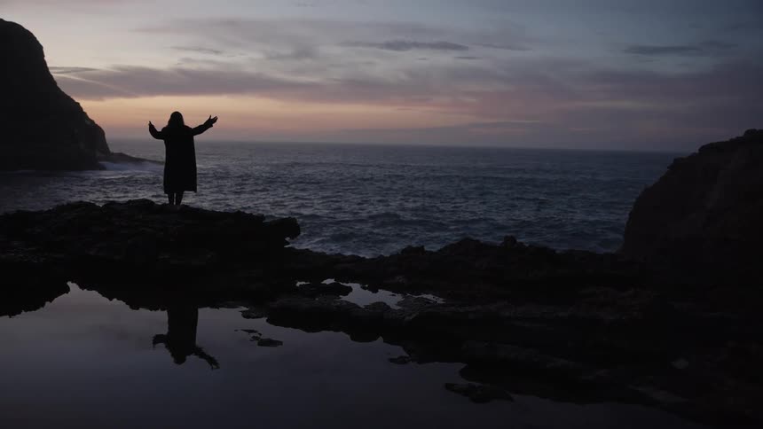 A person stands silhouetted on shore rocks between land formations with arms raised facing the open water under a fading sky their form mirrored in a foreground pool of water