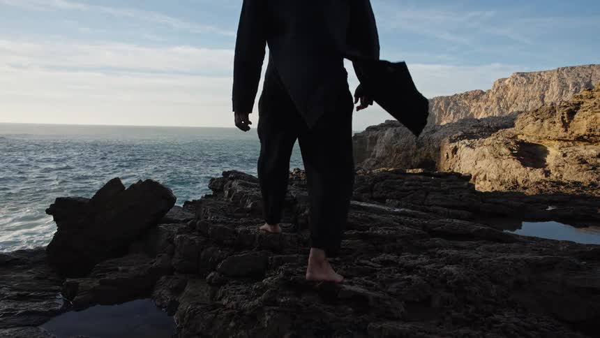 A rear medium view of a barefoot person wearing a long black coat and black pants as they walk carefully across dark jagged rocks by the ocean on a bright day