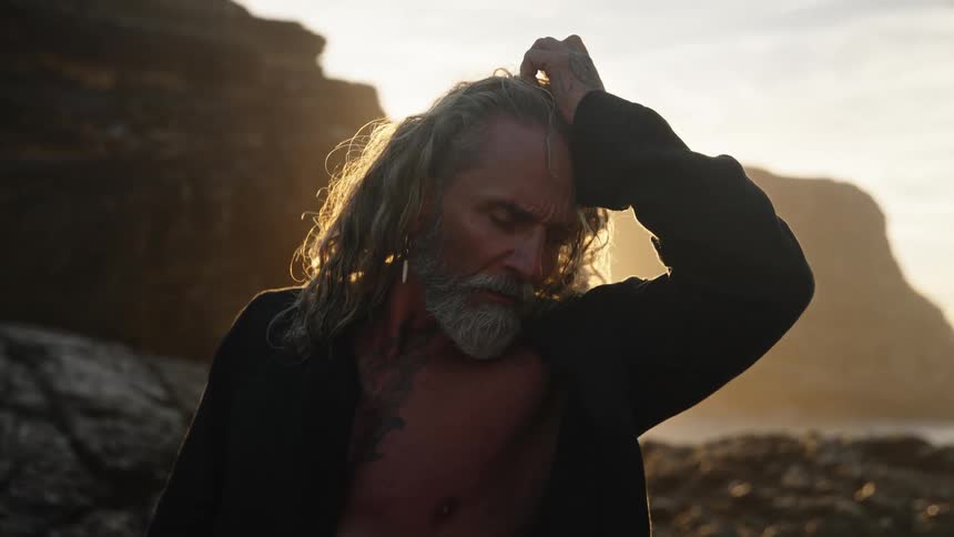Man with grayish hair and beard standing on rocks with golden light in the background