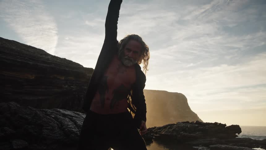 Tattooed man with long hair in expressive pose near rocky shoreline at sunset