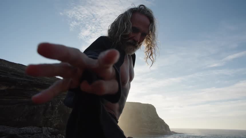 Grayhaired man opening a coat in a coastal landscape at sunset