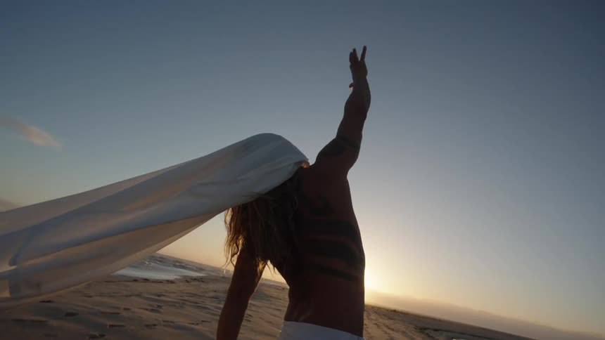 A low angle medium view shows a shirtless tattooed person with long hair wearing white pants standing on a beach with a white sheet covering their head and one arm raised toward the setting sun