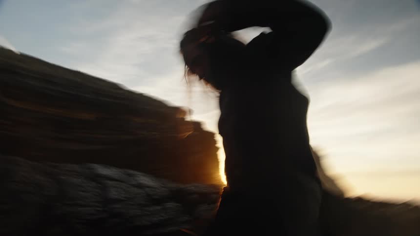 A medium view shows the dark silhouette of a man with long hair wearing a black shirt and pants as he dances across wet rocks into the blinding sun setting behind a cliff face