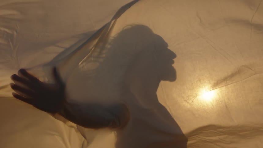 A close view shows the silhouette of a person with long hair touching a translucent light brown sheet that is being dramatically backlit by a bright light source that casts deep shadows