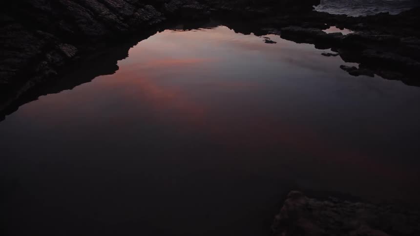 A close view shows the still water of a dark tide pool reflecting the faint colors of the sunset sky surrounded by dark rugged rocks near the ocean