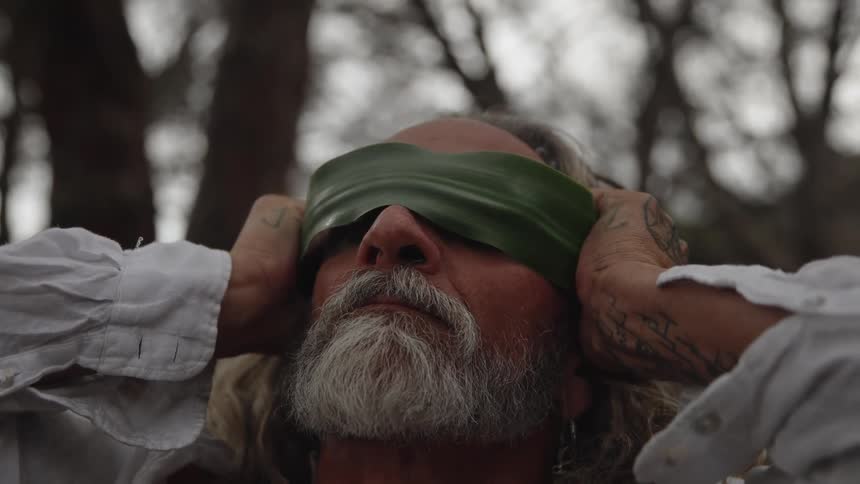 A bearded older man with long hair tied back is shown in a closeup as he adjusts a dark green leaf or fabric blindfold over his eyes outdoors in a blurred forest or garden setting his hands one of which has a dark tattoo on the forearm are vis