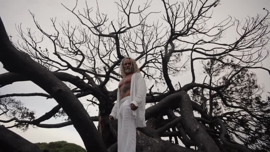 A middleaged man stands on a leafless tree trunk his white shirt unbuttoned to reveal tattoos on his chest paired with trousers