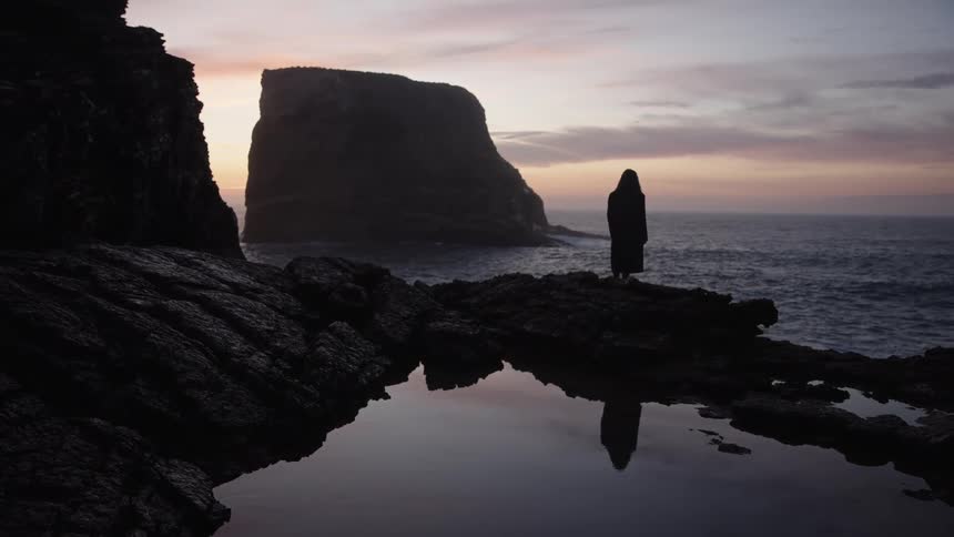 Person in a dark coat watching the sea at sunset reflected in a puddle among rocks