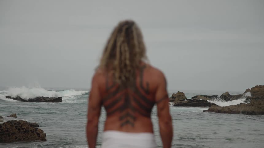 Man with long wavy hair stands on rocks facing the sea displaying symmetrical back tattoo