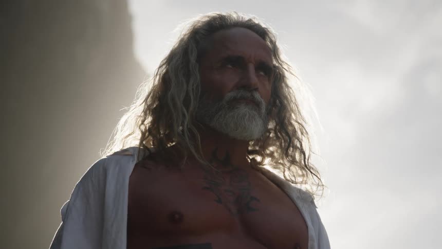 The video is a medium closeup of a man with long hair and a beard shirtless and wearing a white garment draped over his shoulders he is dramatically lit from behind by a strong bright white light that creates a halo effect and washes out the b