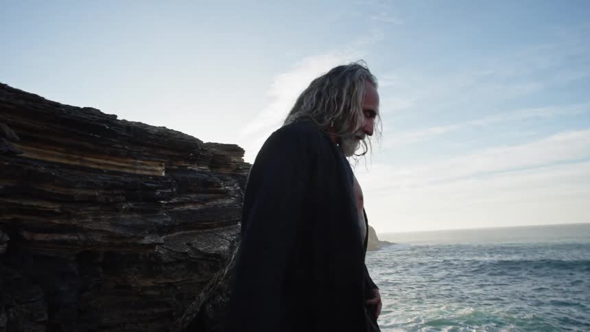 A side medium view of a shirtless older man with long hair a gray beard and chest tattoos wearing a long black coat as he stands on a rocky cliff overlooking the ocean and raises his arms toward the blue sky
