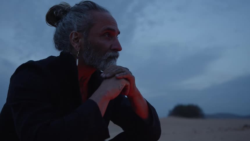 A man with a gray beard and bun is sitting pensively in a desertlike setting at dusk his hands are clasped under his chin as he looks into the distance with a contemplative expression