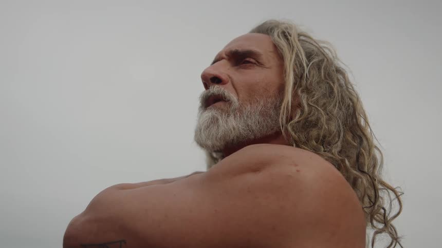 A profile close view of a shirtless older man with long curly blonde hair and a gray beard posing with his arms crossed and looking up against a bright pale sky