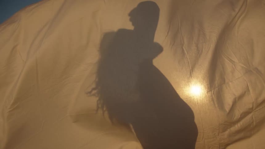 A close view of the silhouette of a person with long hair dancing behind a flowing beige colored sheet illuminated by bright sunlight